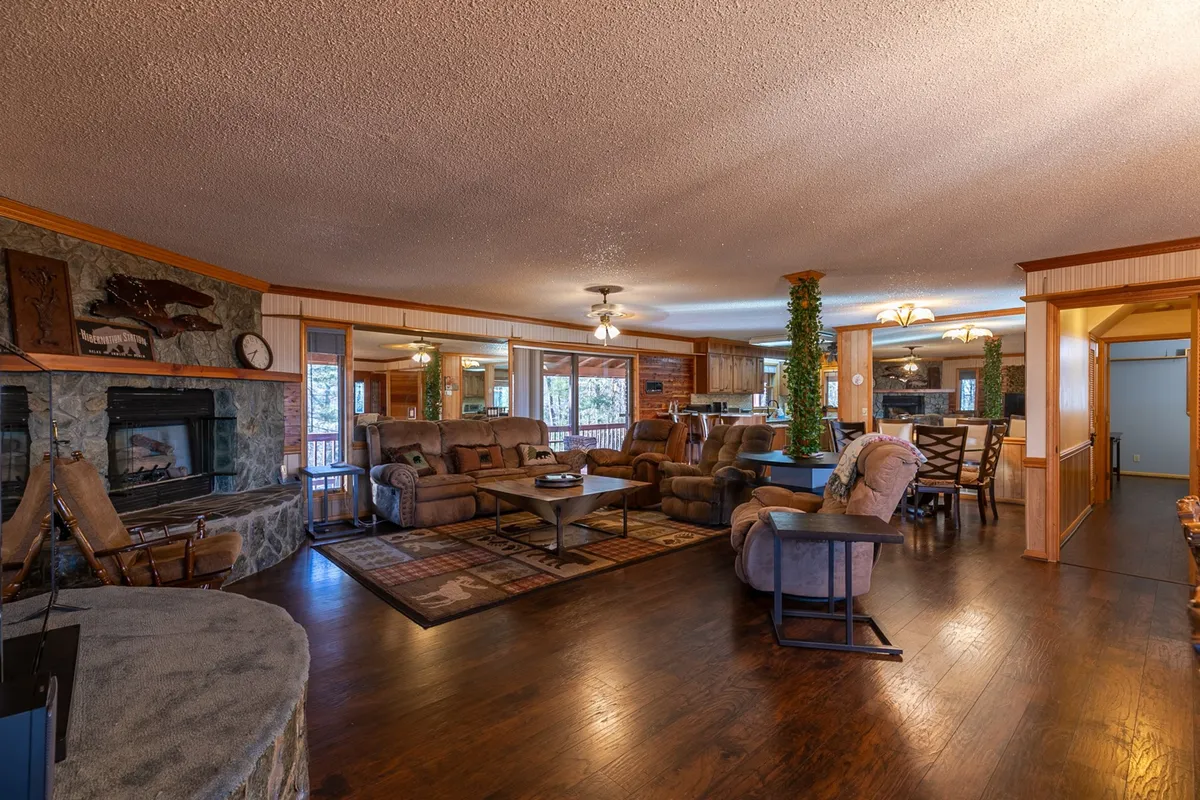 An open-concept living area featuring a large seating arrangement around a flickering gas fireplace, with visible wooden accents and a spacious dining area in the background. The scene is set up for gatherings, cozy relaxation, and family activities, with games on the soft rugs and a table ready for meals.