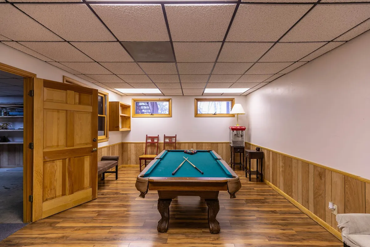 A warmly lit recreation space featuring a pool table, wooden chairs for spectators, and a unique light fixture overhead. The friendly atmosphere suggests a place for both playing and relaxing, suitable for all ages to come together.