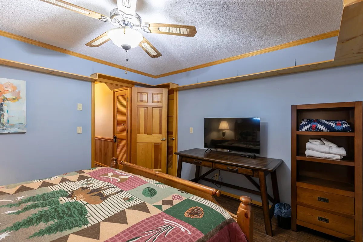 A welcoming bedroom with a bed dressed in a patterned quilt, warm wooden furniture, a television for evening relaxation, and a painting adding a touch of homeliness. The window allows natural light to brighten the space.