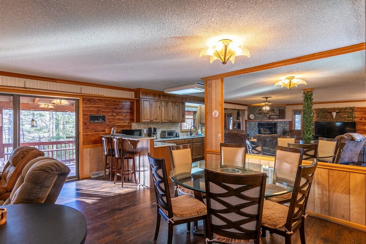 A warm, inviting view of the dining table adjacent to a kitchen island, with ample seating and a comfortable living area nearby. Large windows let in natural light, highlighting the space designed for memorable family dinners, cooking sessions, and cozy evenings.