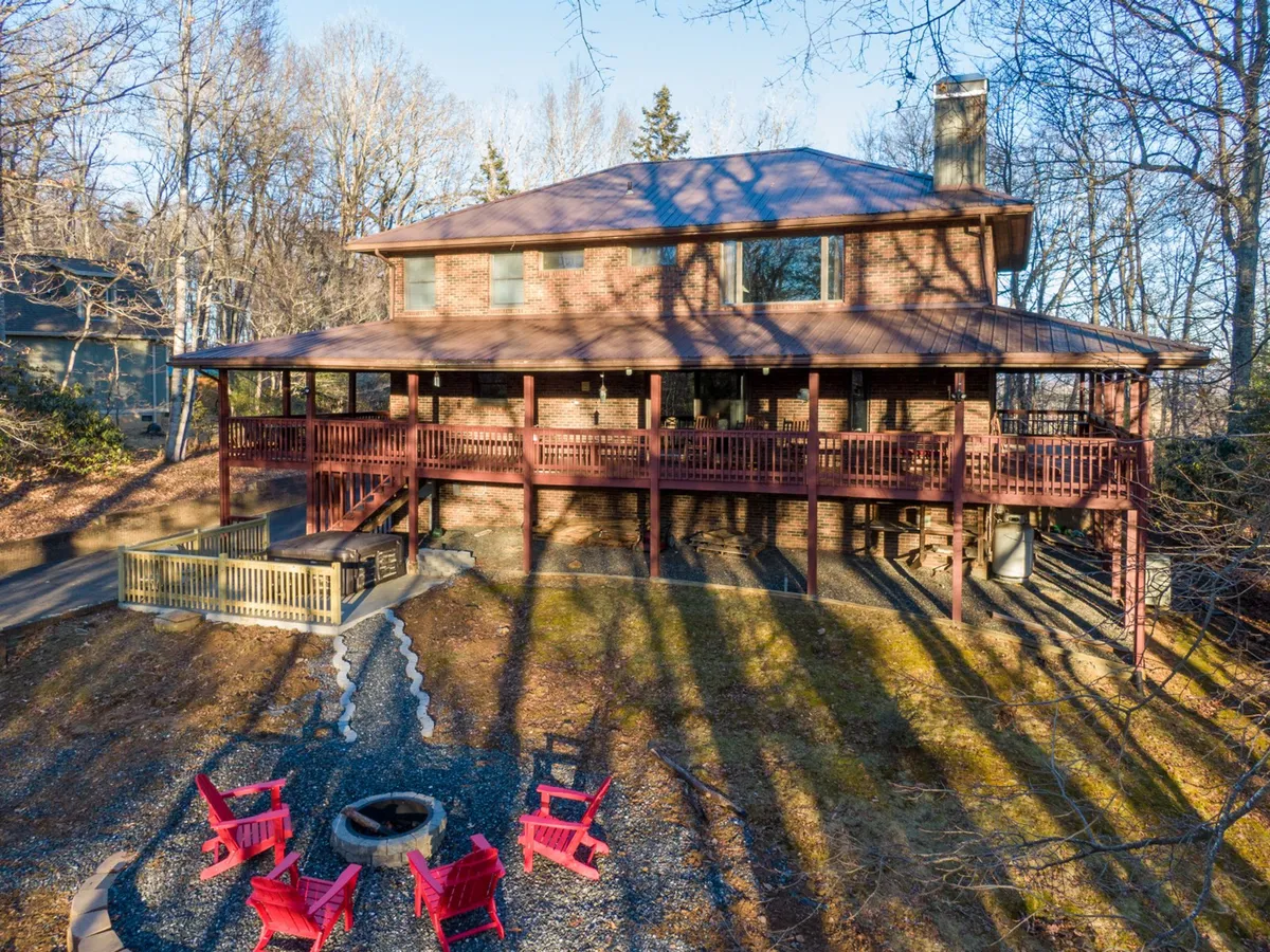 A large wooden deck extends from the brick facade of the house, featuring cheerful red outdoor chairs and a central fire pit. The deck is surrounded by trees and offers ample space for outdoor activities and gatherings.