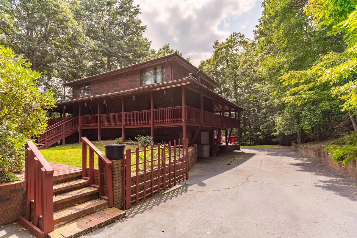 The hero image showcases the charming brick facade of a spacious single-family house set amid lush trees, with a large wooden deck inviting guests to relax. The driveway leads straight to the entrance, and the surrounding natural landscape hints at peaceful seclusion. The deck promises sunset views and space for gatherings, while the wooded backdrop enhances the feeling of a mountain retreat.
