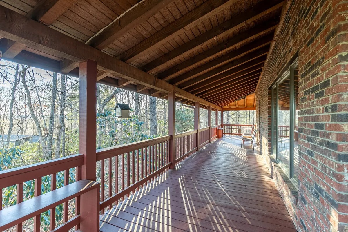 A serene wrap-around deck bordered by a wooden railing, overlooking lush trees and featuring a bird feeder. The deck's flooring and inviting setting evoke a sense of connection with the natural environment and offer ample space for relaxing, enjoying coffee, or letting pets stretch their legs.