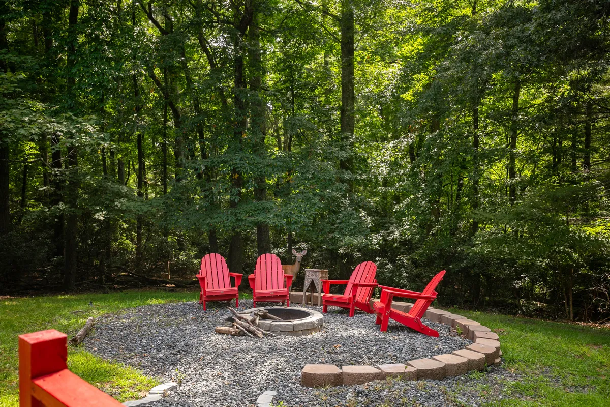 A group of red Adirondack chairs is arranged in a circle around a stone fire pit, surrounded by tall trees and lush greenery. The atmosphere is cozy and welcoming, perfect for campfire gatherings and storytelling under the stars.