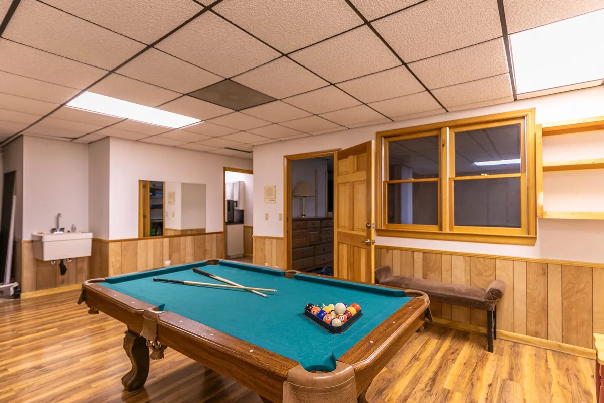 A game room scene with a pool table, wooden shelves filled with games and decor, and built-in bench seating. The setting is casual and communal, highlighting opportunities for both play and conversation.