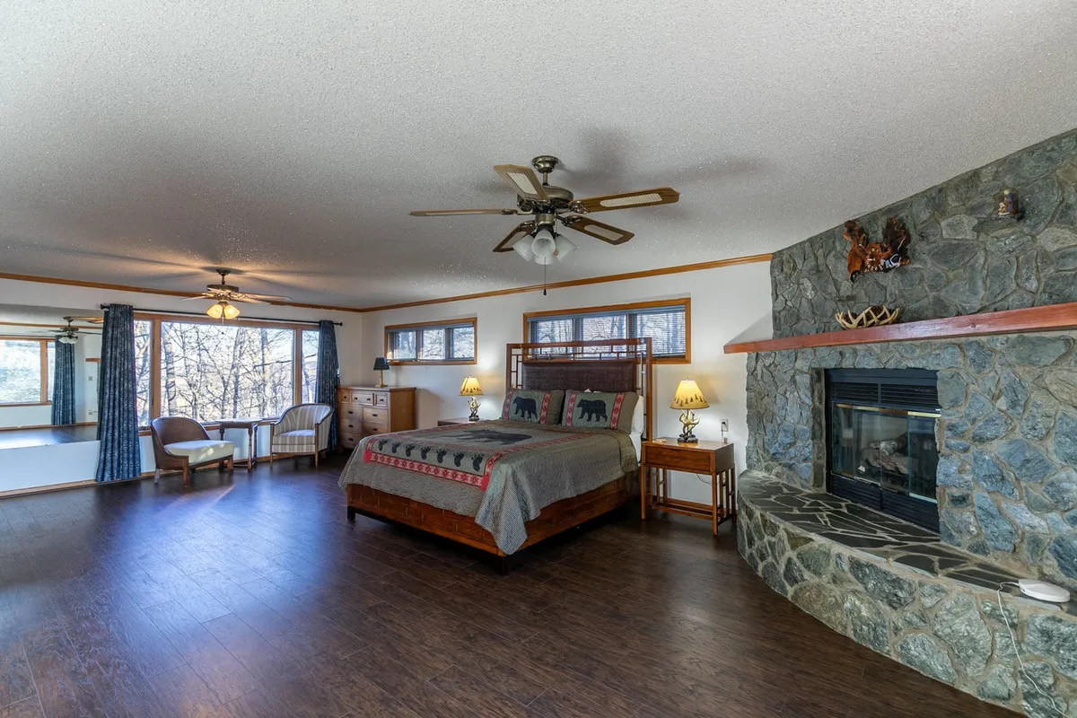 A spacious room featuring a king-sized bed with a decorative blanket, a beautiful stone fireplace, large windows that flood the space with natural light, and a comfortable seating area for relaxing or sharing quiet moments.