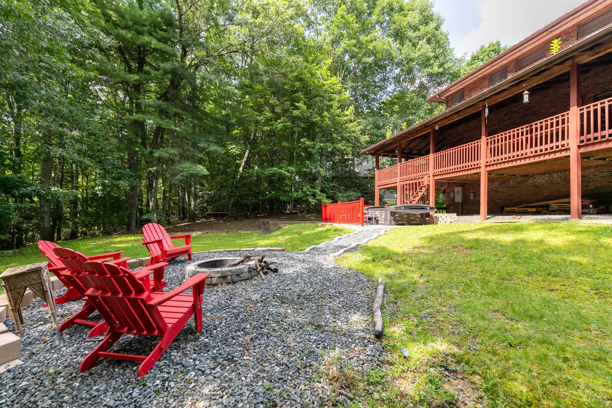 This exterior image captures the backyard setting with red Adirondack chairs encircling a fire pit, a gravel pathway, and lush greenery. The deck of the house can be seen in the background, establishing a direct connection between the deck and the backyard gathering area.