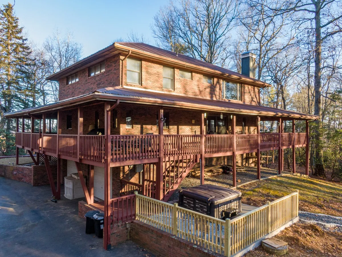 A spacious two-story brick house features a large balcony and an inviting outdoor seating area, complete with a hot tub. The home is set among mature trees, providing a serene natural backdrop. The space is designed for outdoor gatherings, relaxation, and enjoying stunning views.