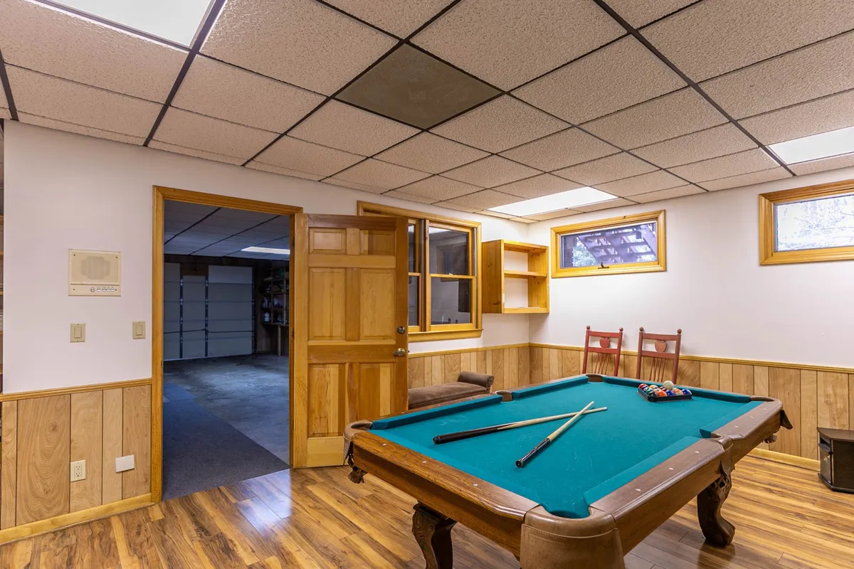 This image captures a dedicated game room with a central pool table, wood paneling, windows letting in natural light, and a wooden door. The space is inviting and clearly intended for recreational activities and socializing, ideal for hosting game nights or friendly competitions.